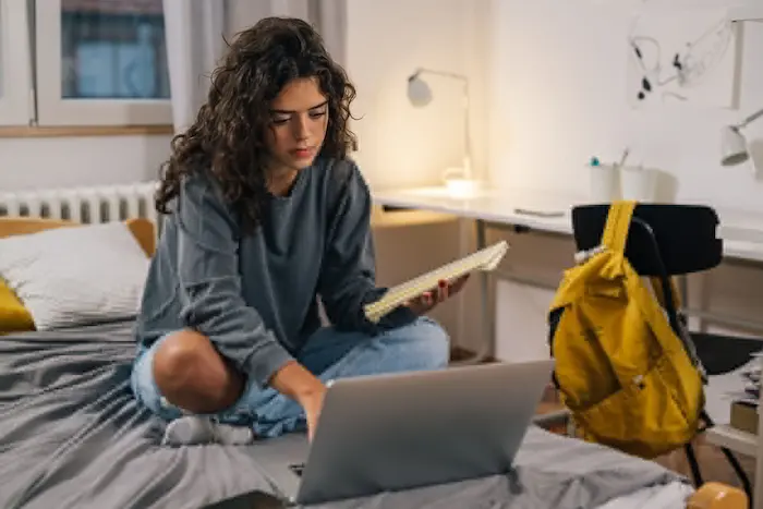 une jeune femme assise sur un lit, en train d'étudier sur son ordinateur, elle est dans la chambre d'une résidence universitaire à Paris